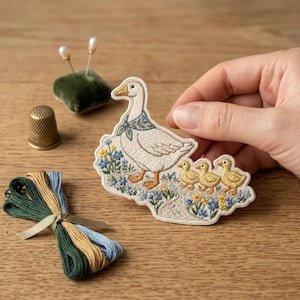 May include: Embroidered patch featuring a white goose with a blue scarf, followed by three yellow ducklings. The patch is surrounded by blue and yellow flowers. Other items include a thimble, pins, and embroidery floss.