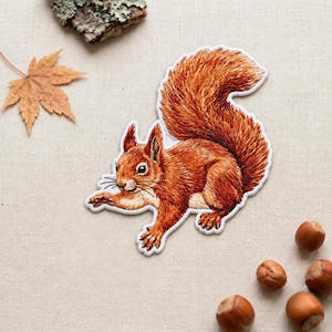 May include: An embroidered patch featuring a detailed red squirrel with a bushy tail, rendered in shades of orange and brown. The squirrel is depicted in a dynamic pose with visible whiskers and textured fur, finished with a white border edge.