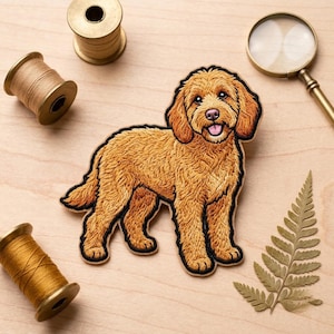 May include: An embroidered patch featuring a golden-brown dog with curly fur, a pink tongue, and dark eyes. The patch is surrounded by two wooden spools of thread, a magnifying glass with a gold handle, and a single dried fern leaf.