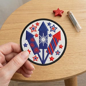 May include: Embroidered patch featuring three stylized fireworks in red and blue, surrounded by red and blue stars. The patch has a black border and is on a white background. A red star and a glass vial are also visible.
