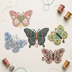 May include: Five embroidered butterfly patches in various colors and designs. Butterflies include floral and geometric patterns, with colors like pink, blue, teal, and yellow. Spools of thread and a thimble are also visible.