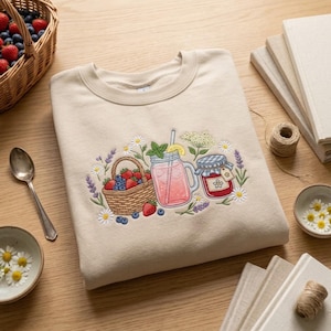 May include: A beige crewneck sweatshirt featuring an embroidered design of a woven basket filled with strawberries and blueberries, a glass jar of pink lemonade with a lemon slice and mint, and a jar of red jam, surrounded by daisies and lavender.