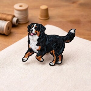 May include: Embroidered patch of a Bernese Mountain Dog in a running pose. The patch features black, white, and brown colors, with detailed stitching. The dog's tongue is visible. The patch is on a white cloth.