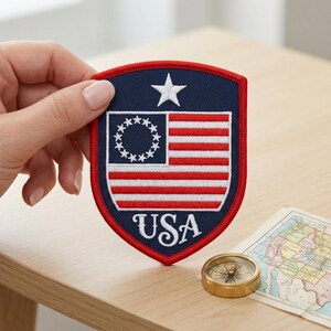 May include: A shield-shaped patch featuring a stylized American flag design with a blue field, red and white stripes, and a white star. The word "USA" is embroidered in white script at the bottom. A small brass compass and a map are also visible.
