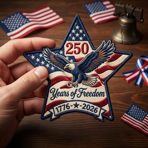 May include: A star-shaped patch with a patriotic design, featuring a bald eagle, the number 250, and the text "Years of Freedom 1776-2026." The patch is in red, white, and blue, with embroidered details. Additional miniature American flags and a bell are in the background.