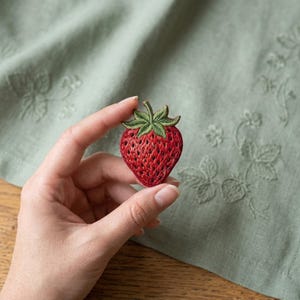 May include: A detailed, embroidered strawberry patch. The patch features a vibrant red body with a textured appearance and a green leafy top. The patch is held in a hand against a soft green background.