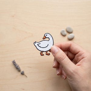 May include: A white goose patch with black outline, orange feet and beak. The patch is held by a hand against a light wood surface. Small, smooth stones and a sprig of lavender are also on the surface.