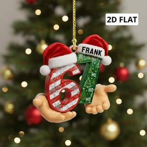 May include: A Christmas ornament shaped like the number 67, held by a pair of hands. The number 6 is red with white stripes and snowflakes. The number 7 is green with a Christmas tree design. Both numbers wear Santa hats. The name "FRANK" is on the number 7.