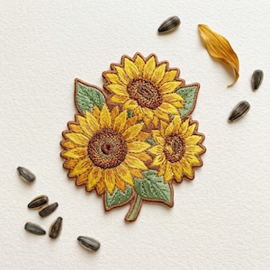 May include: Embroidered patch featuring three sunflowers with yellow petals and brown centers, surrounded by green leaves. The patch is bordered with brown thread. Several sunflower seeds and a dried petal are scattered around the patch.