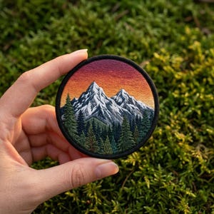 May include: Embroidered round patch featuring a mountain range with snow-capped peaks and evergreen trees. The sky transitions from orange to purple. The patch has a black border and is held against a green, mossy background.