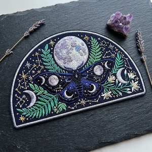 May include: Embroidered patch featuring a moth with a full moon, crescent moons, and stars on a navy blue background. The moth has blue and black wings. Green leaves and white flowers are also included in the design. A purple amethyst crystal and lavender sprigs are also present.