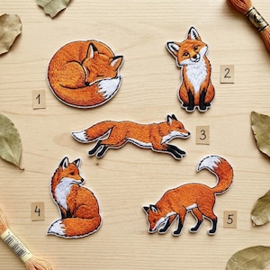 May include: Five embroidered iron-on patches featuring orange and white foxes in various poses, labeled with numbers 1 through 5. The designs include a sleeping fox, a sitting fox, a running fox, a fox looking back, and a foraging fox.