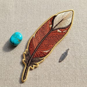 May include: An embroidered feather patch with brown, tan, and gold colors. A small silver feather pendant and a turquoise stone are also present. The patch has detailed stitching and a decorative border.