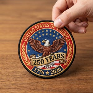 May include: A round embroidered patch featuring a bald eagle with outstretched wings, a banner reading "250 YEARS", and the dates "1776" and "2026". The patch is bordered by the words "UNITED STATES OF AMERICA" and stars.
