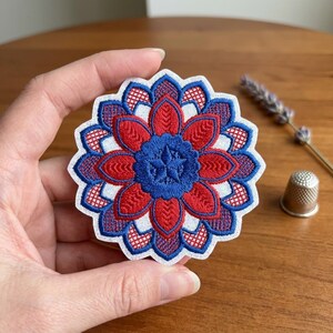 May include: A detailed, embroidered patch in a floral design. The patch features a central blue starburst surrounded by red and blue petals, with white accents. The patch is held in a hand, with a thimble and lavender sprig in the background.