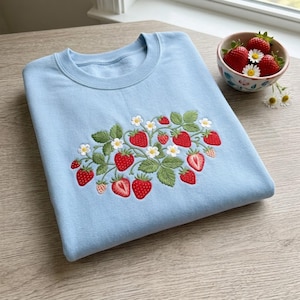 May include: A light blue crewneck sweatshirt featuring an embroidered design of red strawberries, green leaves, and small white flowers. The embroidery is centered on the chest, showcasing various whole and halved strawberries with detailed textures.