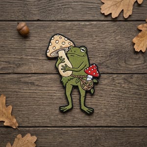 May include: An embroidered patch featuring a green frog standing upright. The frog holds a large, tan, spotted mushroom cap and carries a small woven bag containing a red mushroom with white spots. The design has a black stitched border.