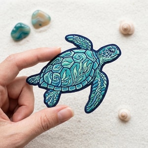 May include: Embroidered patch of a sea turtle in shades of teal and blue, with intricate detailing. The patch is held against a sandy background with small stones and a seashell.
