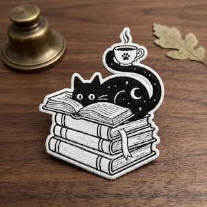 May include: A black and white embroidered patch featuring a black cat with a crescent moon and stars on its body, resting on a stack of three books. The cat's long tail curls upward to hold a steaming cup of coffee with a paw print design.