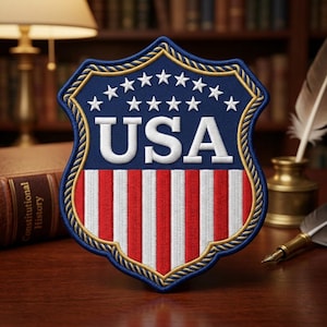 May include: Embroidered shield patch featuring the letters "USA" in white, set against a blue background with white stars. The lower portion displays red and white stripes, all framed by a gold rope-like border. A patriotic emblem.