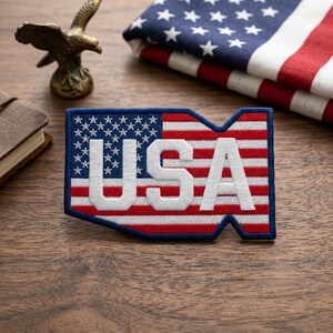 May include: Embroidered patch in the shape of the USA, featuring the American flag design with the word "USA" in white. The patch is on a wooden surface, with a folded American flag and a bronze eagle statue in the background.