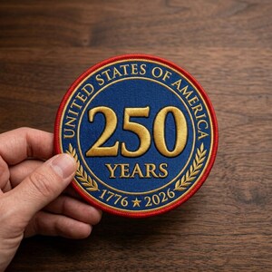 May include: A round patch with a blue center featuring the text "250 YEARS" in gold, surrounded by "UNITED STATES OF AMERICA" and the years "1776 * 2026". The patch has a red border and is held by a hand.