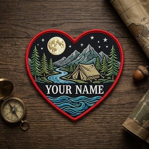 May include: A heart-shaped embroidered patch featuring a night landscape with a full moon, stars, mountains, pine trees, a winding river, and a tent. The text "YOUR NAME" is stitched in white in the center of the patch, which has a red border.