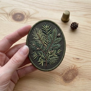 May include: An oval-shaped embroidered patch featuring a detailed design of green pine needles and small brown pinecones on a dark olive green fabric background, with a matching dark green stitched border edge.