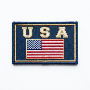 May include: Embroidered patch featuring the American flag and the word "USA" in cream-colored letters. The patch has a navy blue background with a gold border. The flag design includes red and white stripes and a field of white stars on a blue background.