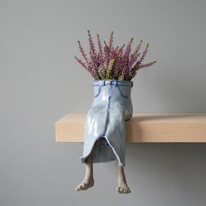 May include: A light blue ceramic vase shaped like a pair of jeans, with legs and feet. The vase is filled with purple flowers. The vase is sitting on a wooden shelf against a gray background.
