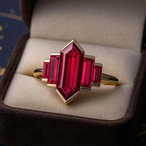May include: A gold ring with a large, rectangular, faceted red gemstone at the center, flanked by smaller rectangular red stones. The ring is set in a gold band and presented in a brown jewelry box.