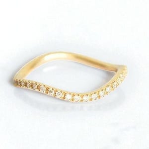 Puede incluir: Un anillo dorado con un diseño ondulado. El anillo está adornado con pequeñas gemas brillantes a lo largo del borde curvo. Se presenta sobre un fondo blanco, resaltando su elegante forma.