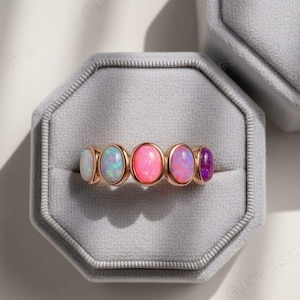 Puede incluir: Un anillo de oro rosa con cinco piedras preciosas ovaladas en fila. Las piedras son blancas, azules, rosas, moradas y violetas. El anillo se muestra en un joyero de terciopelo gris.