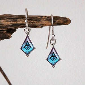 May include: A pair of silver-toned drop earrings. Each earring features a square-cut, teal-colored gemstone with a geometric design. The gemstones are set in a silver metal frame and hang from a simple hook.