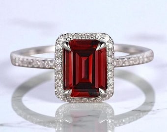 May include: A silver ring featuring a rectangular, deep red gemstone, surrounded by a halo of smaller, clear stones. The band is also adorned with small, clear stones. The ring is displayed on a reflective surface.