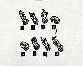 May include: A white t-shirt with a black graphic of hands signing the word "FILM NOIR". The text "PKON KAVE" is printed above the graphic.