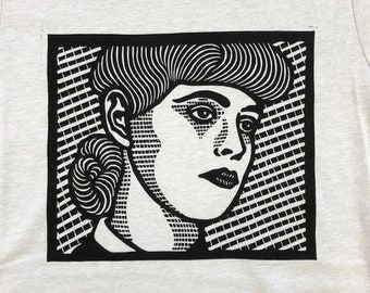 May include: A white t-shirt with a black and white graphic print of a woman's face. The graphic is in a square frame with a black and white checkered pattern.