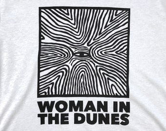 May include: A white t-shirt with a black and white graphic of a stylized eye surrounded by lines. The text "WOMAN IN THE DUNES" is printed below the graphic.