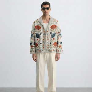 Puede incluir: Chaqueta color crema con intrincados bordados florales en rojo, azul y verde. La chaqueta tiene un cuello a rayas y se combina con pantalones color crema. El conjunto se completa con gafas de sol oscuras y sandalias.