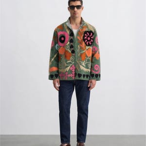 Puede incluir: Una chaqueta verde con bordados florales coloridos, incluyendo diseños rosas, naranjas y negros. La chaqueta tiene cremallera y se combina con vaqueros azul oscuro y mocasines de cuero marrón. El diseño de la chaqueta incluye corazones y otras formas.