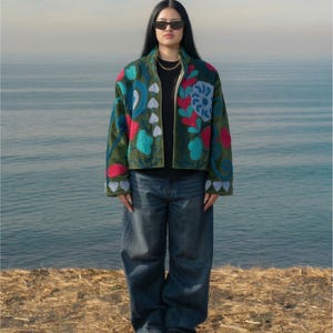 Op de afbeelding: Een groene jas met kleurrijke bloemenborduursels, gedragen met een zwarte top en een blauwe jeans. De jas heeft rode, blauwe en witte bloemmotieven. De persoon draagt een zonnebril en een gouden ketting.