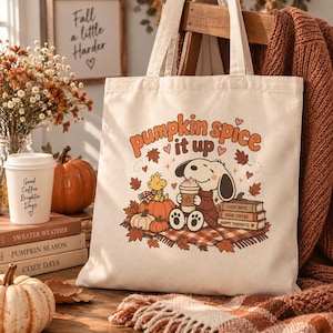 May include: A cream-colored canvas tote bag featuring a graphic of Snoopy and Woodstock sitting on a plaid blanket with pumpkins, autumn leaves, and a stack of books. The text reads pumpkin spice it up, cozy days, good coffee, and happy thoughts.