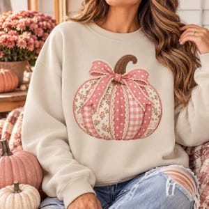 May include: A cream-coloured crewneck sweatshirt featuring a large central graphic of a pumpkin. The pumpkin is composed of vertical panels in various pink patterns, including floral, polka dot, and gingham, accented with a pink polka dot bow.