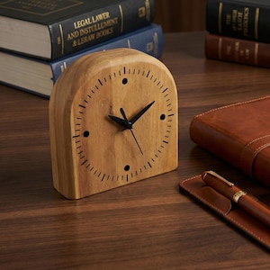 Peut inclure: Une horloge de bureau en bois avec des aiguilles noires et un design minimaliste. L'horloge est en bois clair et repose sur une surface en bois, à côté d'un journal en cuir, d'un stylo et de livres.