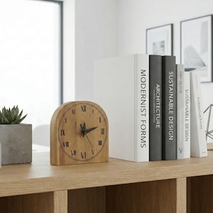 May include: A wooden clock with Roman numerals, a succulent plant in a concrete pot, and a stack of books with titles like "Modernist Forms" and "Sustainable Design" are arranged on a wooden shelf. A geometric metal sculpture is also present.