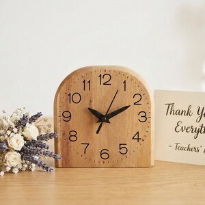 May include: A wooden desk clock with black hands and numerals, next to a small bouquet of flowers and a card that reads "Thank You for Everything - Teachers' Day". The clock is approximately 13 cm tall.