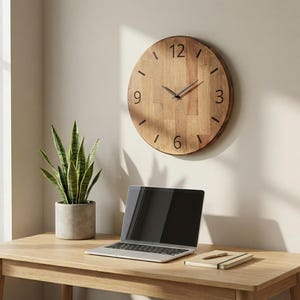 May include: A round wooden wall clock with black numerals and hands hangs above a desk. A laptop, a small notebook, and a potted snake plant sit on the light-colored wooden desk. The clock is approximately 12 inches in diameter.