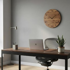 May include: A modern home office setup featuring a wooden desk with a laptop, a potted plant, and a wooden wall clock. The desk has a black metal frame. The clock is round and made of wood. A gray office chair is in front of the desk.