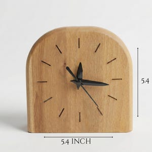 Peut inclure: Une horloge de bureau en bois avec une finition marron clair. Le cadran de l'horloge a des aiguilles noires et des repères linéaires simples. L'horloge mesure 13,7 cm de large et 13,7 cm de haut. Le dessus de l'horloge est arrondi.