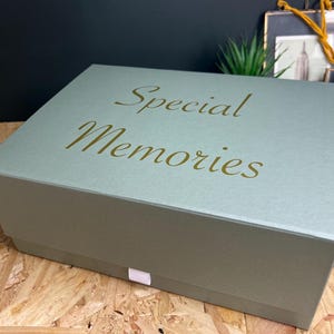 May include: A sage green rectangular box with the words "Special Memories" in gold script on the lid. The box is on a wooden surface. A small white tab is on the front of the box.